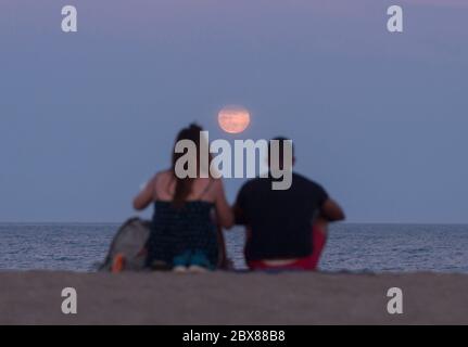 Malaga, Spanien. Juni 2020. Der Erdbeervollmond, als Teil einer Mondfinsternis, steigt über den Horizont, während die Menschen das gute Wetter am Strand von Malagueta genießen, inmitten der Lockerung der Coronavirus-Krankheit (COVID-19) Beschränkungen. In diesem Monat Juni sind die Erdbeer-Vollmond- und Halbmondfinsternis am 5. Juni das erste astronomische Ereignis, das die Menschen sehen können. Während dieses Monats wird auch eine Sonnenringfinsternis am 21. Juni zu sehen. Quelle: SOPA Images Limited/Alamy Live News Stockfoto