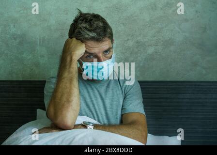 Dramatische Porträt von kranken Mann trägt medizinische Gesichtsmaske auf dem Bett sitzen Angst und besorgt infiziert durch Coronavirus leiden Lockdown allein zu Hause p Stockfoto