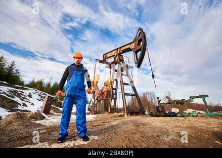 Ölarbeiter während seines Arbeitstages auf dem Ölfeld, Mann mit blauen Overalls und orangefarbenem Helm, hält Rohrschlüssel, Blick auf die Kamera unter dem schönen Himmel. Konzept der Ölförderung, Erdölindustrie. Stockfoto