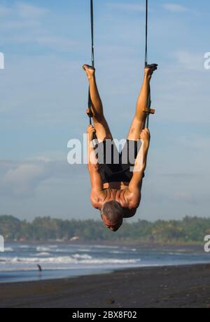 Outdoor-Porträt des Mannes üben Aero Yoga Training am Strand hängen von Seil Schaukel über dem Meer Training Körperbalance und Flexibilität in der hea Stockfoto