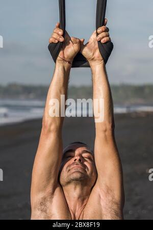 Outdoor-Porträt des Mannes üben Aero Yoga Training am Strand hängen von Seil Schaukel über dem Meer Training Körperbalance und Flexibilität in der hea Stockfoto