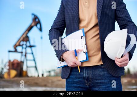 Ausschnitt-Schnappschuss eines Mannes Ingenieur hält seine Notiz und einen Stift in der einen Hand und einen weißen Helm in der anderen, im Ölfeld stehend, Ölpumpenheber ist auf Hintergrund, Konzept des Ölgeschäfts Stockfoto