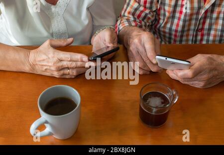 Draufsicht auf alte Menschen, die Smartphones zu Hause benutzen, während sie Tassen heiße Getränke genießen. Konzept der älteren Menschen und Technologie. Stockfoto