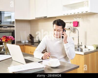 Junge asiatische Geschäftsmann sitzen Küche Zähler arbeiten zu Hause sprechen auf Handy Notizen Stockfoto