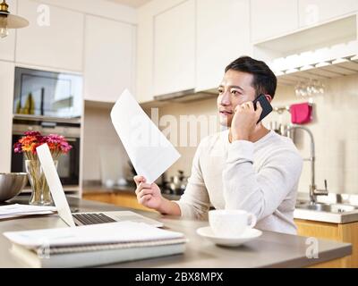 Junge asiatische Geschäftsmann sitzen Küche Zähler zu Hause arbeiten sprechen auf Handy halten ein Dokument Stockfoto