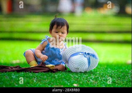 Isoliert offen Porträt von süß und liebenswert asiatisch koreanisch Baby Mädchen 3 oder 4 Monate alt spielt mit Hut allein im Stadtpark sitzen auf grünem Gras in Stockfoto