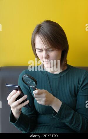 Ein junges, schönes Mädchen von europäischem Aussehen in einer blauen Jacke sitzt in einem Café mit einem Handy auf der Couch vor dem Hintergrund einer gelben Wand. Schaut durch eine Lupe auf die Gadgets-Anzeige. Stockfoto