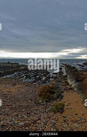 Felsschichten, die in das Meer bei St Monan's an der Fife Coast tauchen, neben dem äußeren Wellenbrecher des kleinen Hafens, mit dunklen Winterwolken darüber. Stockfoto