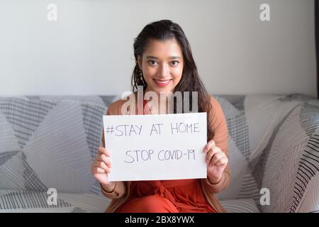 Glückliche junge schöne indische Frau zeigt Aufenthalt zu Hause Zeichen, während unter Quarantäne aufgrund von Covid-19 Pandemie Stockfoto