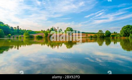 Romantische Landschaft mit kleiner Kirche auf dem Hügel spiegelt sich im Teich. Sonniger Sommertag mit blauem Himmel und weißen Wolken. St. Peter und Pauls Kirche in Bysicky bei Lazne Belohrad, Tschechische Republik. Stockfoto