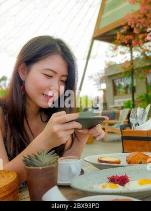 Junge hübsche und glücklich asiatische koreanische Reise-Blogger Frau, die Bild mit Handy der schönen Frühstückstisch im Freien Café lächelnd Stockfoto