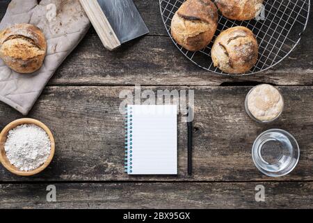 Draufsicht des leeren Rezept Notizblock mit schwarzem Marker auf rustikalem Holztisch platziert von hausgemachten Sauerteig Brot Brötchen und Zutaten umgeben. Stockfoto