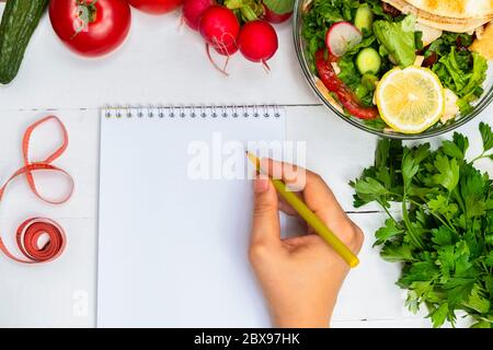 Draufsicht der leeren weißen Notizblock und Frau Hand halten Stift auf weißem Holztisch mit gesundem Salat und frischem Gemüse. Gesundes Diät-Konzept Stockfoto