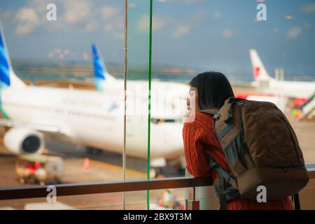 Junge glückliche und aufgeregt asiatische chinesische Studentin mit Rucksack am Flughafen Abflug Lounge beobachten Flugzeuge durch Glasfenster lächelnd bereit für Stockfoto