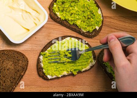 Holzbrett mit Avocado-Sandwiches, Brot und einer offenen Packung Frischkäse. Der Prozess der Herstellung von Toast mit Avocado. Vegetarisches Gericht. Ein Blick von oben Stockfoto