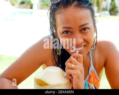 Lifestyle Naturportrait der 30er oder 40er Jahre glückliche und attraktive asiatische Indonesierin im Bikini, die Kokoswasser am tropischen Resort Swimmingpool trinkt