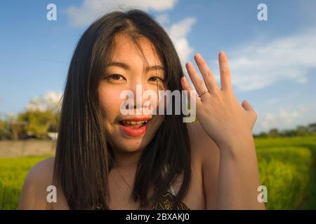 Junge glückliche und schöne asiatische Frau im Freien zeigt stolz ihren Verlobungsring. Fröhliche chinesische Mädchen aufgeregt als Verlobter lächelt auf grünes Feld Stockfoto