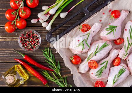 Rohe Hähnchenschenkel auf einem Backblech auf Pergament mit Rosmarin und Gewürzen, frisches Gemüse mit Olivenöl auf Holzhintergrund, Draufsicht Stockfoto