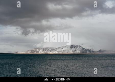 Ein schneebedeckter Berg an der Atlantikküste Islands. Blick von der anderen Seite oder vom Meer. Wolkig, dichte Wolken. Stockfoto