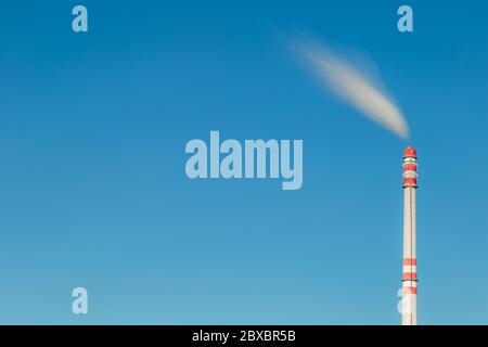 Industrieller Schornstein im Wind. Schmutziger Rauch aus rot-weißem Fabrikschornstein am blauen Himmel. Lange Belichtung, Platz für Text. Stockfoto