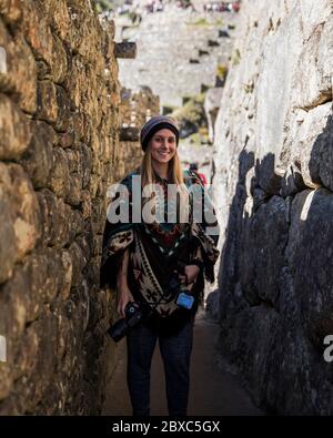 Blonde junge Frau, die zwischen zwei Steinwänden mit einer Kamera in der Hand und einem peruanischen Outfit steht. Stockfoto