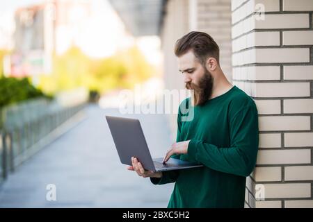 Ein schöner bärtiger Mann, der einen Laptop in der Straße der Stadt hält und benutzt Stockfoto