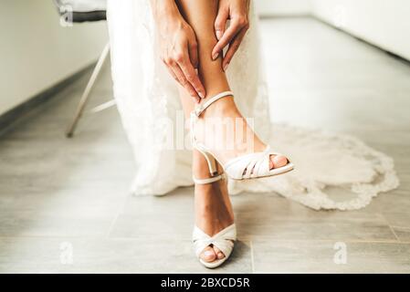 Braut setzt weiße Schuhe an ihren zarten Füßen. Frau im Hochzeitskleid sitzt auf Stuhl - Beine aus nächster Nähe. Stockfoto