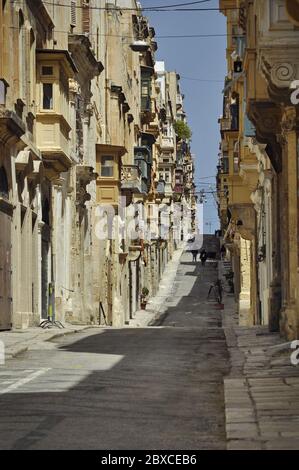Blick auf die Straße bergauf mit hellen Steinhäusern auf beiden Seiten der Straße mit maltesischen Balkonen. Fußgänger sind im Hintergrund Stockfoto
