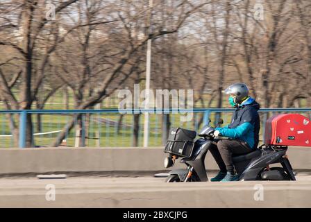 Belgrad, Serbien - 20. März 2020: Reifer Mann mit Sturzhelm, Gesichtsmaske und Handschuhen, Roller mit schwarzer Tasche im Vorderteil und roter Heckauflage Stockfoto