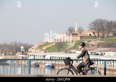 Belgrad, Serbien - 19. März 2020: Junge Frau mit Fahrradhelm, die über die Stadtbrücke mit der Festung Kalemegdan im Hintergrund fährt Stockfoto