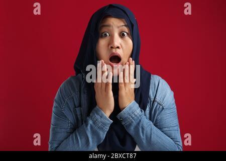 Schockiert überrascht asiatische muslimische Dame Blick auf Kamera mit großen Augen und offenen Mund, wow betäubt Geste über roten Hintergrund Stockfoto
