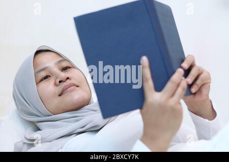 Asiatische muslimische Frau, die den heiligen Koran auf dem Bett liest, um den Koran vor dem Schlaf auswendig zu lernen, islamische Kultur Stockfoto