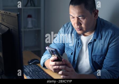 Asiatischer Mann mit Telefon-Anwendung und arbeitet auf seinem Computer in der Nacht, Online-Zahlungsbestätigung Stockfoto