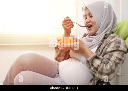 Glücklich asiatische muslim schwangere Mutter sitzt auf Liegestuhl und essen ihre gesunde Nahrung für ihr Baby am Nachmittag Stockfoto