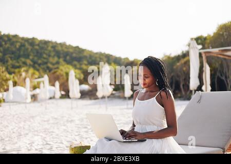 Porträt von lächelnden hübsche junge Frau in weißem Sommerkleid sitzt auf Chaiselongue und arbeiten auf Laptop Stockfoto