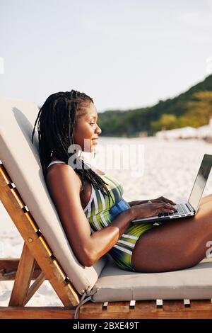 Lächelnd hübsche junge schwarze Frau im Badeanzug sitzt auf Chaiselongue und arbeiten auf Laptop Stockfoto