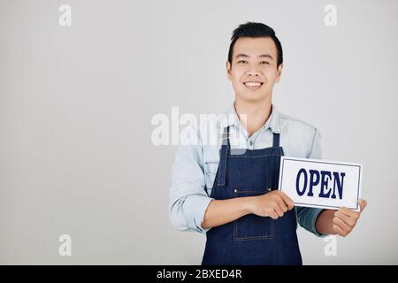 Glückliche junge Cafe-Besitzer in Denim Schürze hält offenes Zeichen, isoliert auf grau Stockfoto