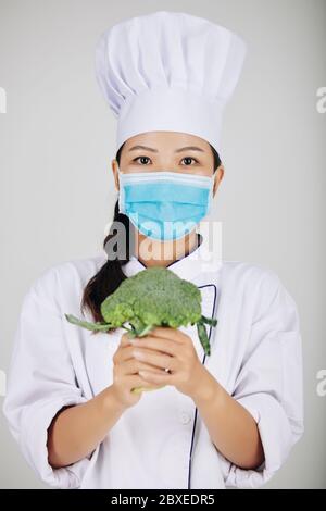 Junge weibliche Cafe Chef in medizinischen Maske hält frischen Brokkoli Stockfoto