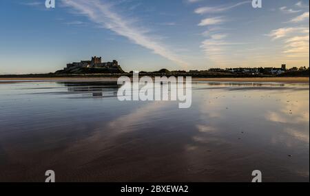 Bamburgh Castle fotografiert vom Strand in warmen Abendsonnen und blauem Himmel mit hohen Zirruswolken, die sich alle in einer weiten Weite von nassem Sand spiegeln Stockfoto
