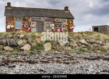 Kabine am Meer aus rostigen Metallwänden, Asbestdach, mit Segelzubehör dekoriert, auf einer grasbewachsenen Bank an einem Kiesstrand unter einem bewölkten Himmel Stockfoto