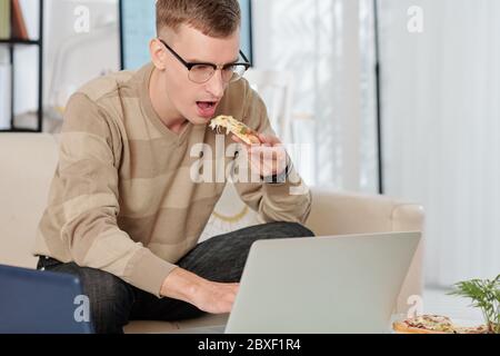 Junger Mann in Gläsern, der eine Pizza isst, wenn er E-Mails von Kollegen auf dem Laptop liest Stockfoto