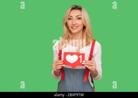 Social Media Blog gefällt und folgt ihm. Porträt von schönen lächelnden erwachsenen Frau in stilvollen Denim Overalls halten Netzwerk Herz-Symbol, Emoji Benachrichtigung aber Stockfoto