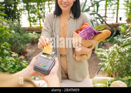 Nahaufnahme einer lächelnden Dame, die einen Papiertüten voller Gemüse hält und mit Kreditkarte für Bio-Lebensmittel im Supermarkt bezahlt Stockfoto