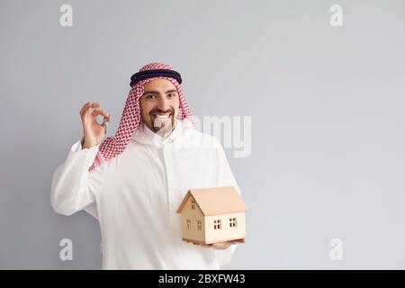 Arabischer Mann makler mit Holzhausmodell in der Hand auf grauem Hintergrund. Stockfoto