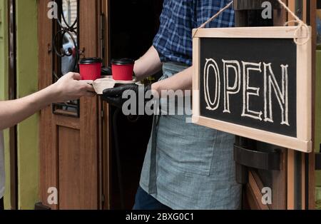 Die Hände des Kellners halten zwei Tassen Kaffee in der Nähe der Tür des Cafés. Arbeiter gibt einen Auftrag zum Mitnehmen für Kunden des Straßenmarktes. Stockfoto