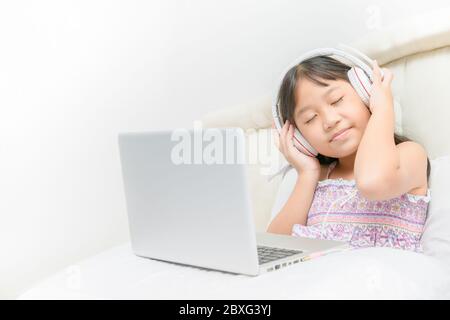 Nette glückliche asiatische Mädchen tragen Kopfhörer, um Musik von Laptop auf dem Bett im Schlafzimmer hören. Bildung und Technologie-Konzept Stockfoto