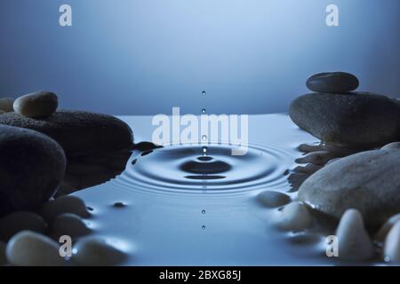Perfekter Wassertropfen in einem Teich mit Wellen. Blaue Töne, um Pfütze in der Nacht von Kieselsteinen umgeben reflektieren. Friedliche Ruhe. Stockfoto