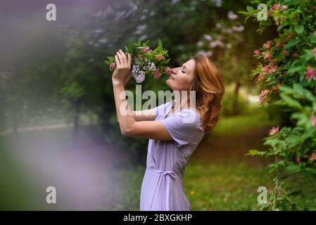 Frau in lila Kleid schniffs Blumenstrauß mit geschlossenen Augen und verschwommenen Frühling bunten Hintergrund Stockfoto