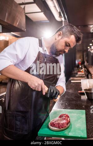 Ein ernster bärtiger Koch, der ein schürzenpeppierendes Steak serviert, während er es in der Küche des Restaurants mariniert Stockfoto