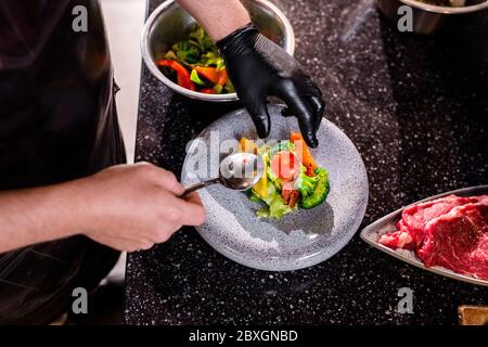 Blick über den unerkennbaren Koch in Latexhandschuh, der das Essen auf den Teller legt, während er das Gericht für Restaurantgäste zubereitet Stockfoto